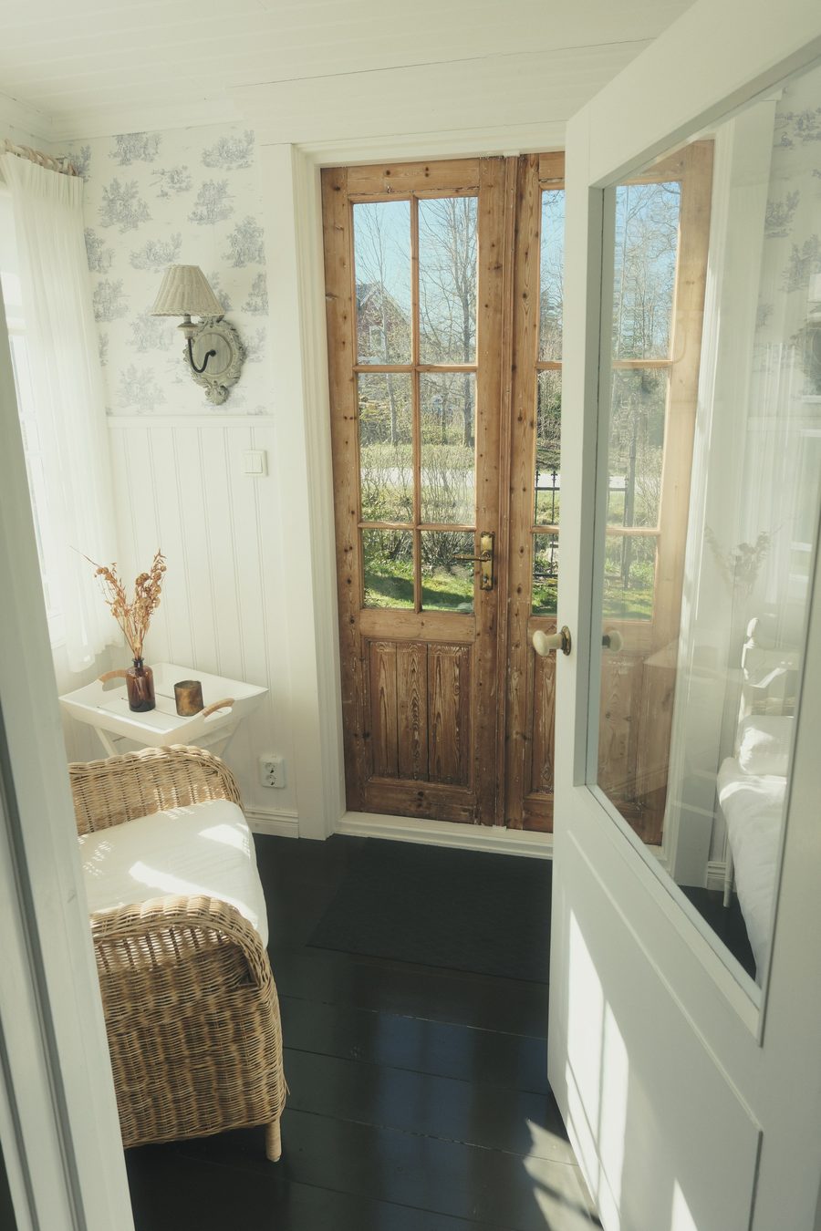 Sunlit entryway with blue toile wallpaper above white beadboard, a wicker armchair and glazed wooden doors