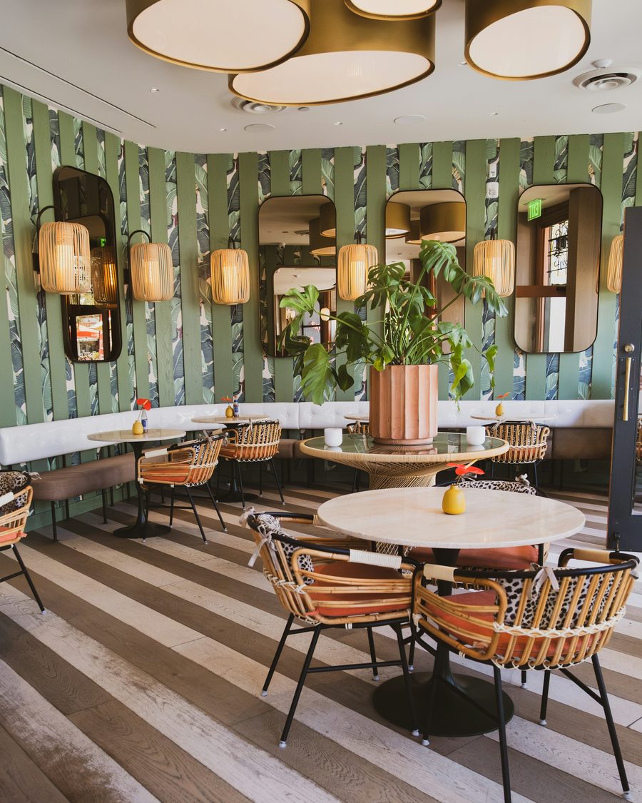 Boutique café with banana-leaf wallpaper behind sage wood slats, rounded mirrors, woven lanterns and rattan chairs
