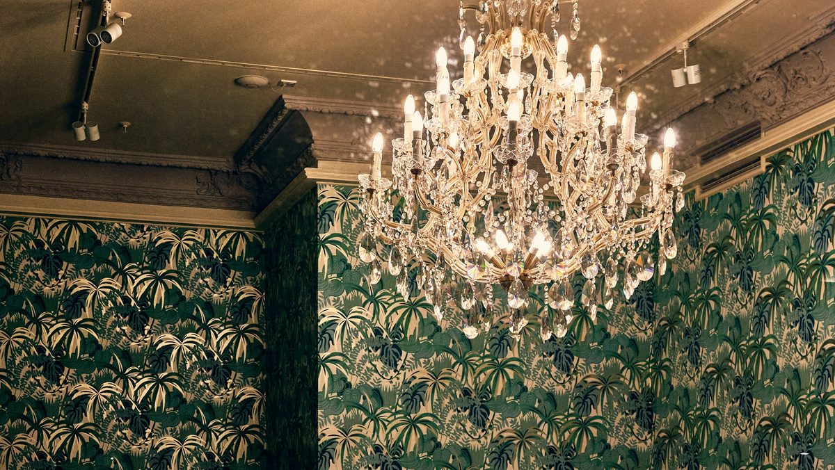 Intimate dining room lined with deep green palm-print wallpaper beneath a lit crystal chandelier
